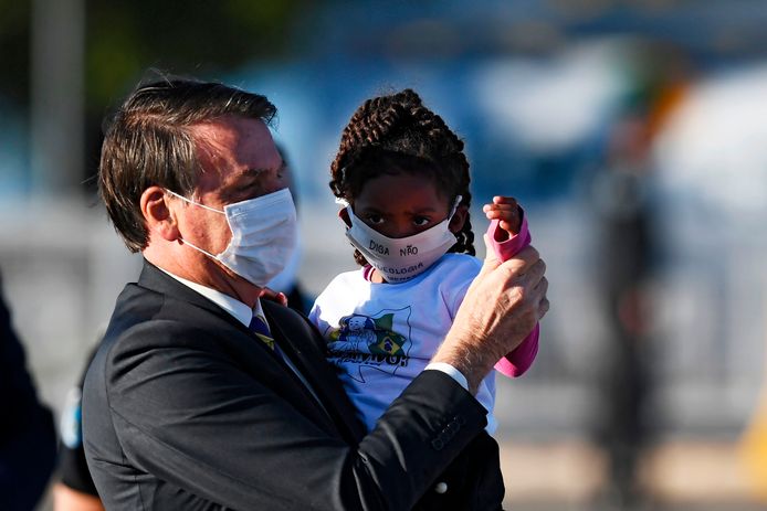 De Braziliaanse president Jair Bolsonaro tilt een meisje op. Beide dragen een mondkapje.