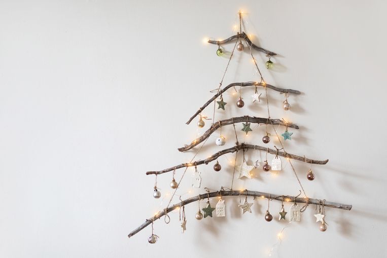 Handmade rustic Christmas tree made from branches with glowing garland lights and ceramic ornaments hanging on white wall. DIY inpiration. Christmas and New Year concept. Beeld Getty Images