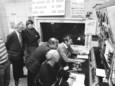 Heren die in 1990 belangstellend naar een panel met knoppen en wijzertjes kijken. Misschien zeggen de gezichten of borden aan de wand jou iets?