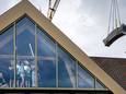Een grote kraan hijst de betonnen bank over het dak van cultureel centrum De Voorste Venne in Drunen. Een aantal kinderen kijkt vanuit het cultureel centrum toe.