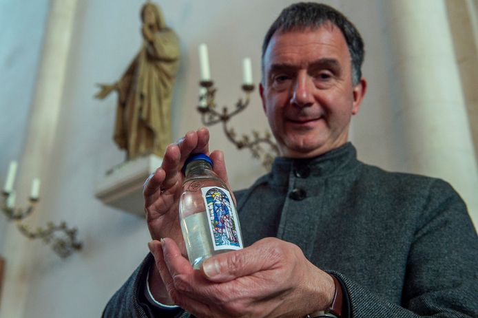 Pastoor Paul verbeek toont een flesje gevuld met heilig Gertrudiswater.  Op de achtergrond is het beeld van de heilige St. Gertrudis te zien.