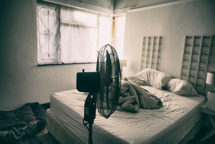 Electric fan in-front of an unmade bed with light coming through a window