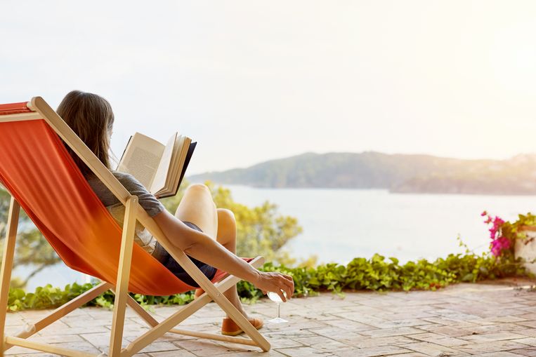 Woman reading book while relaxing on deck chair at back yard Beeld Getty Images