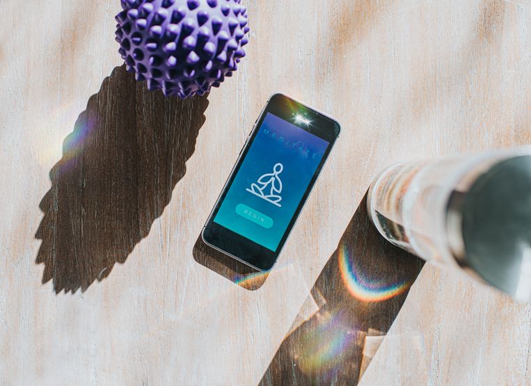 Flat lay image of a mobile phone displaying a meditation app. Beside it is a crystal water bottle and a purple spiked massage ball, casting a shadow. Space for copy. Beeld Getty Images