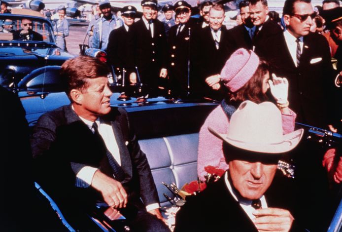 The president and his wife in the back of a limousine leaving Love Field Airport in Dallas.  In the foreground he wears a governor's hat.