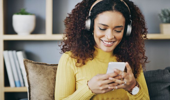 Shot of a young woman wearing headphones while using her cellphone at home