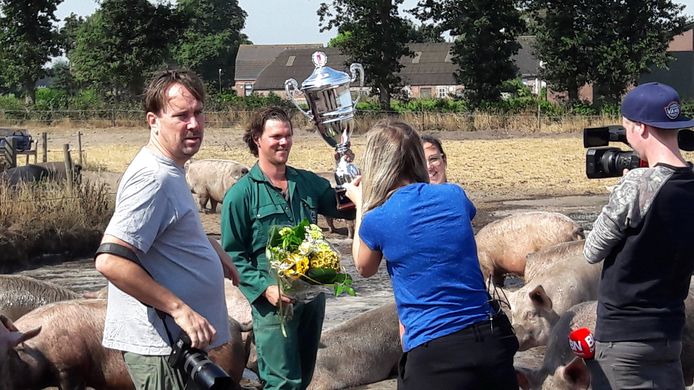 Boer Bas Antonissen toont de Modderpoeltrofee aan de verzamelde media, terwijl achter hem de varkens genieten van de modderpoel.