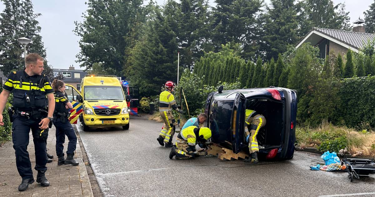 Auto op zijn kant na botsing met boom, automobilist kruipt gewond uit wagen.