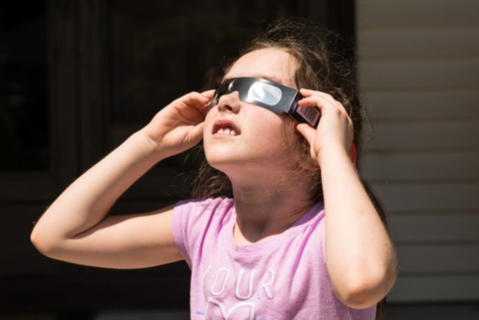 Een meisje draagt een eclipsbrilletje om naar een zonsverduistering te kijken. Zo'n bril is nodig om je ogen te beschermen.
