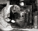 Kolenkachel. Vrouw in ochtendjas hurkt naast de  haard en prikt met een pook in de brandende kolen, om het vuur op te stoken. Op de kachel een waterketel en ernaast een koperen kolenkit.