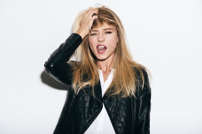 Funky young woman in leather coat holding hand in hair and sticking out tongue while standing against white background