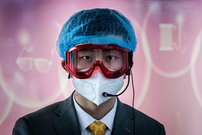Een werknemer bemant een informatiedesk op de internationale luchthaven van Beijing.  Hij draagt een beschermende bril,  haarkapje en mondmasker.
