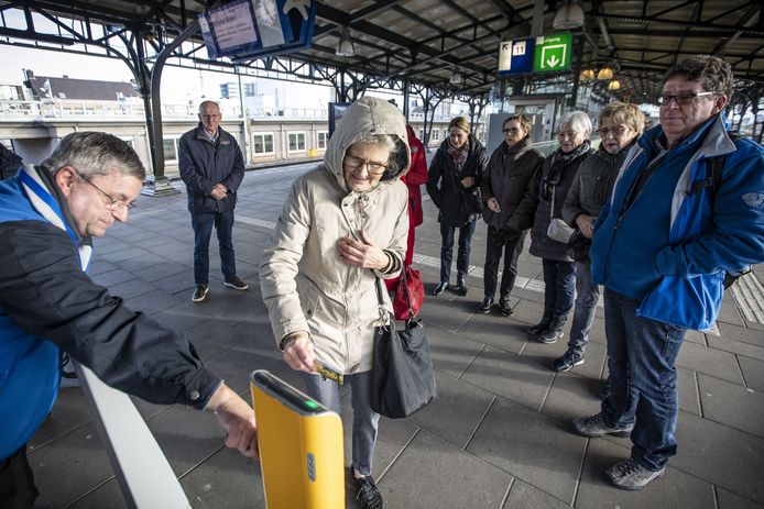 De 75-jarige Astrid (midden) houdt haar OV-chipkaart tegen de incheckpaal, op aanwijzen van OV-ambassadeur Arjen Mengerink (links).