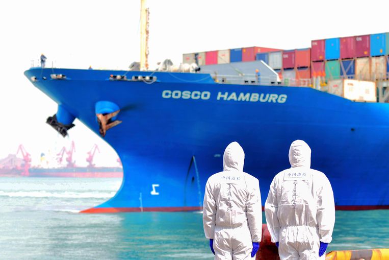 Werknemers in beschermende pakken kijken naar een containerschip in de haven van de Chinese stad Qingdao. Beeld AP
