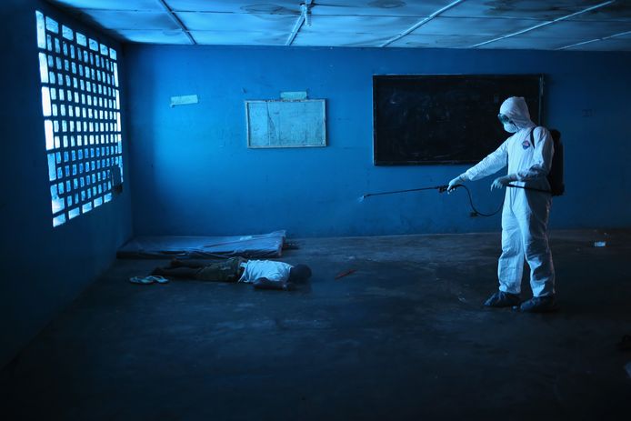Een Liberiaanse gezondheidswerker ontsmet in een klaslokaal het lichaam van een man die aan ebola is overleden. De WHO kwam bij de bestrijding van de ebola-uitbraak in 2014 door politiek geharrewar pas laat in actie.