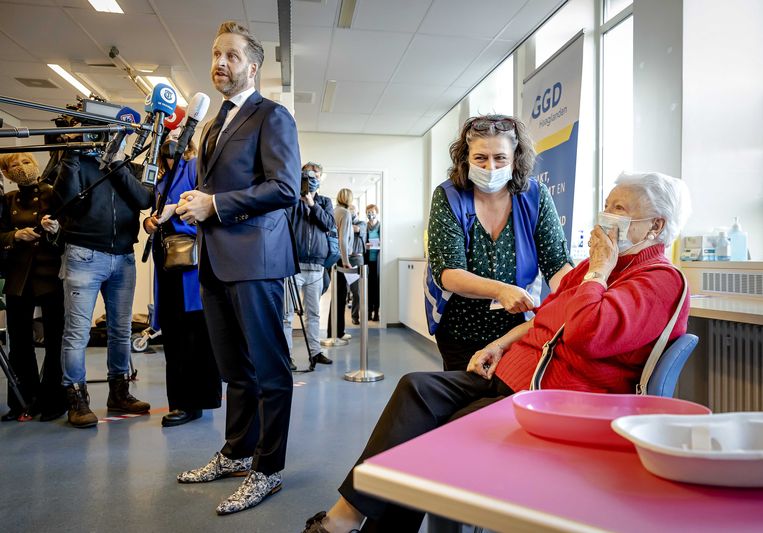 Demissionair Minister Hugo de Jonge van Volksgezondheid, Welzijn en Sport (CDA) spreekt met de pers terwijl een GGD-medewerker een boosterprik zet bij een oudere inwoner van Den Haag.  Beeld ANP