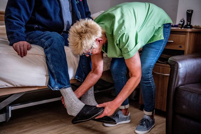 Een medewerkster helpt een oudere man uit zijn schoenen op zijn kamer in een verzorgingstehuis.