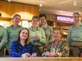 Cliënten van SDW kijken toe terwijl bestuurders Roos Herman van Bibliotheek West-Brabant (links) en Ingrid van Huijkelom van SDW de samenwerkingsovereenkomst bezegelen.