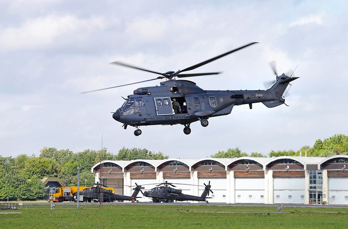Een cougar helikopter vliegt langs de flightline van vliegbasis Gilze-Rijen. Op de achtergrond de hangaars met ervoor geparkeerde apaches.