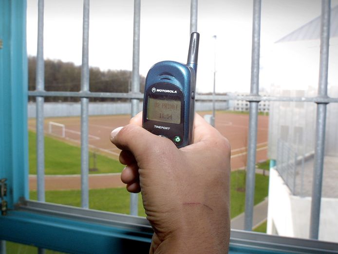 Een man houdt een oud type mobiele telefoon vast terwijl hij achter de tralies zit.