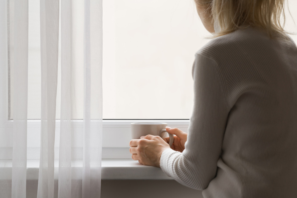 Sad adult woman sitting at window and holding mug in hands. Looking out from home. Thinking about life. Back view. Beeld Shutterstock
