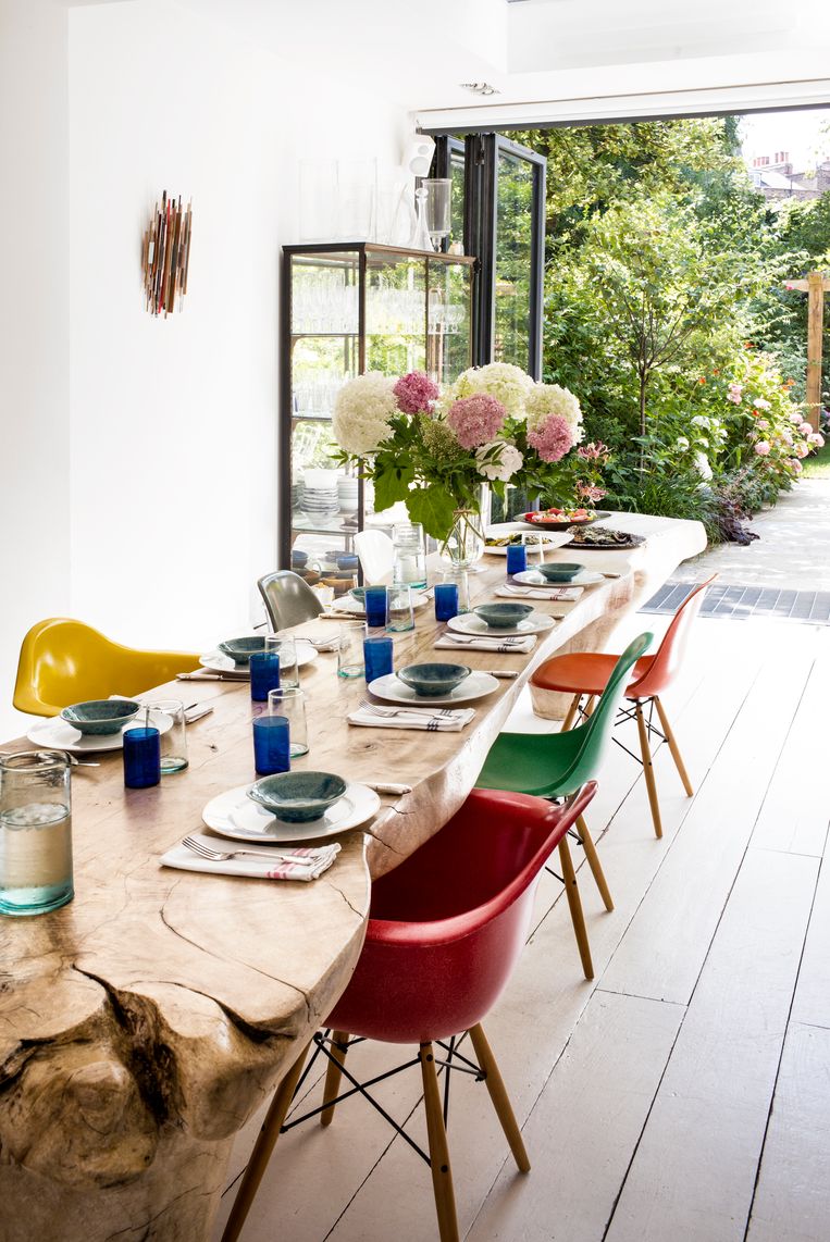 Kleurige Eames-stoelen omgeven het pronkstuk van het huis, een boomstronktafel uit Israël. De vaas is gevuld met pioenen en hortensia’s uit de tuin. Beeld Paul Raeside