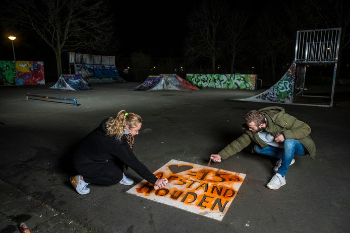 Straathoekwerkers Caroline en Jesse spuiten een boodschap op de grond met oranje haarverf: Liefde is... 1,5 meter afstand. De klus wordt in de avond in het donker gedaan om te voorkomen dat groepjes komen kijken wat ze aan het doen zijn.