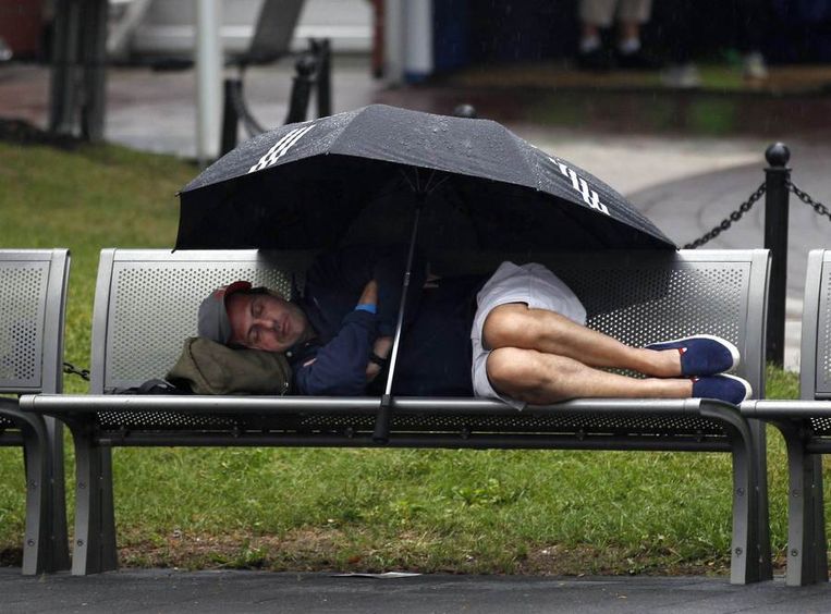 Een bezoeker van de US Open zoekt dinsdag zijn toevlucht onder een paraplu op een bankje. Beeld reuters