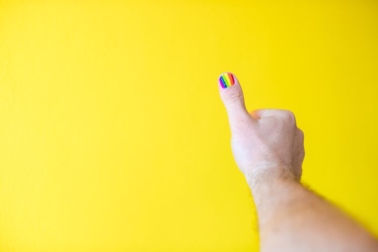Isolated thumb up sign with a rainbow flag on the nail against a bright yellow background - POV picture Beeld Getty Images/iStockphoto