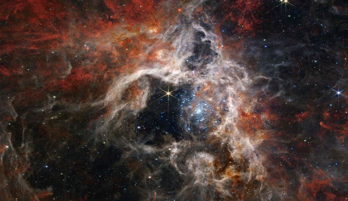 De gaswolk of nevel ligt in het Grote Magelhaense Wolk-sterrenstelsel, dat zich 161.000 lichtjaren (één lichtjaar is ongeveer gelijk aan 9.460.000.000.000 kilometer, red.) van onze Aarde bevindt en in een baan rond de Melkweg draait. De nevel wordt ook wel ‘30 Doradus’ genoemd en is een gebied met duizenden sterren, meer bepaald de heetste en grootste sterren die astronomen kennen. 

De fonkelende blauwe sterren in het midden van de foto hebben de kracht om het gas om zich heen weg te blazen en zijn dus verantwoordelijk voor de holte in de nevel. Rond de middelste holte zijn de stoffige vezels te zien waaruit het ‘web van de Tarantula-nevel’ bestaat.

Alleen de meest compacte gebieden rond de nevel kunnen aan de kracht van de sterren weerstaan, en vormen zo zones waarin nog meer jonge sterren, of ‘protosterren’, worden geproduceerd. Wanneer die sterren ‘volgroeid’ zijn, komen ze uit hun cocon en geven ze de nevel op hun beurt vorm.

De felle ster in het midden is op zich niet speciaal, maar is wel erg helder waardoor de zogenaamde ‘diffractiepieken’ te zien zijn. Die pieken zijn het gevolg van de spiegels van de James Webb-telescoop en de manier waarop die het licht naar de wetenschappelijke instrumenten sturen.